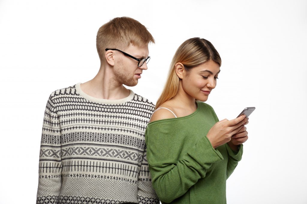 People, relationships and modern technology. Handsome unshaven young male in glasses having distrustful look while suspecting his girlfriend of cheating on him, reading messages on her cell phone